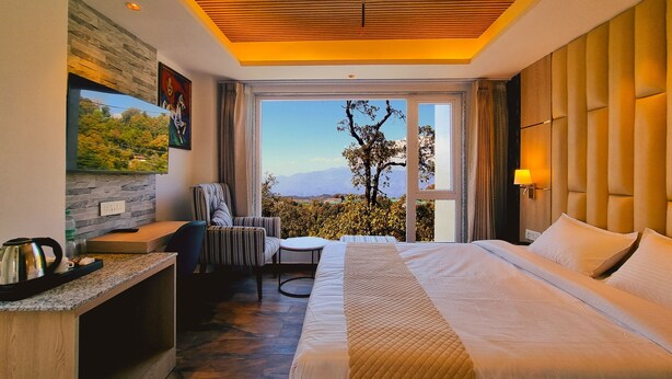 Una habitación de hotel moderna con una cama grande, un escritorio con hervidor, una silla, una mesita y un ventanal amplio con vistas panorámicas.
