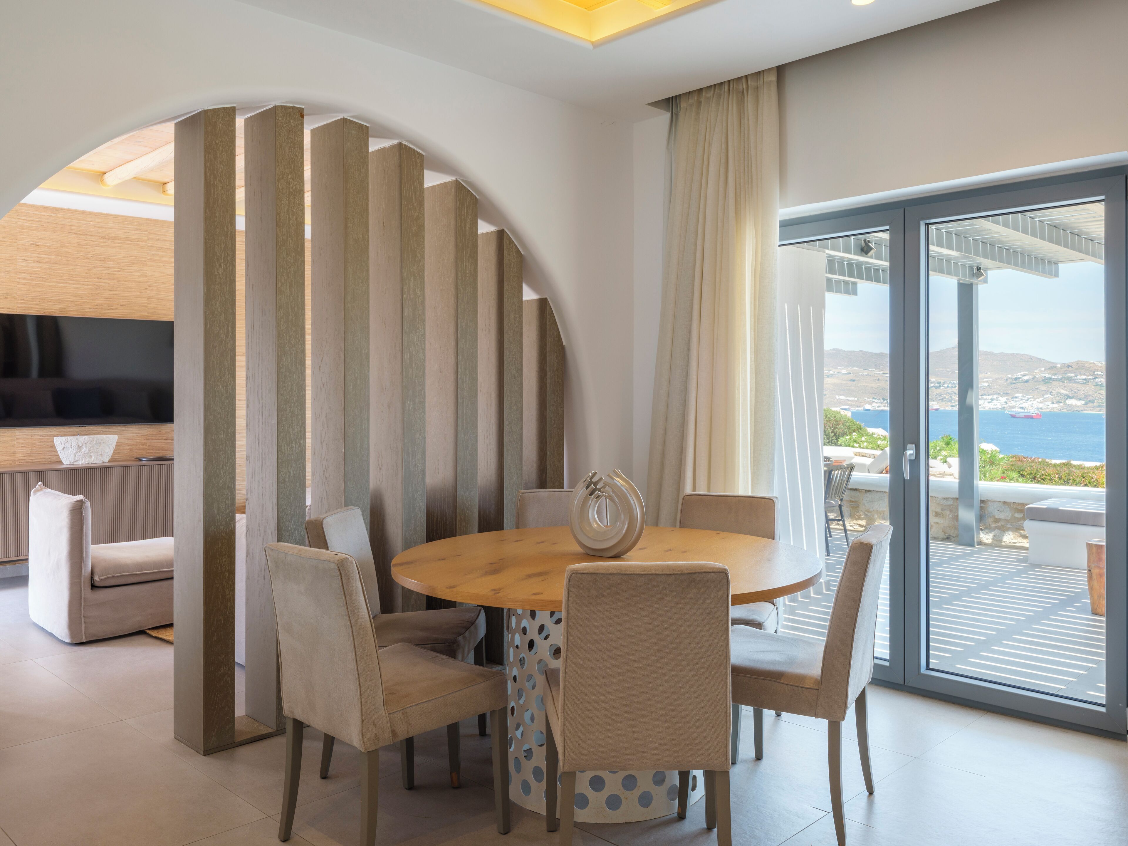 Ein modernes Esszimmer mit einem runden Tisch, sechs Stühlen und Meerblick durch große Glastüren.