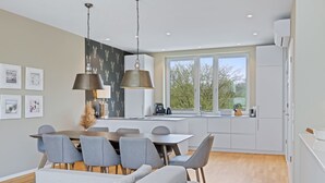 A modern kitchen with a dining area, featuring a large window, pendant lights, and a neutral color palette.