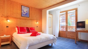 A hotel room with a wooden ceiling, a bed with white and red bedding, a television on a wooden table, and a window with a view of buildings. - Hotel Blanche Neige   (Courchevel)