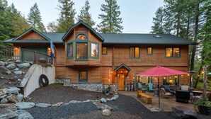 A log cabin with a stone retaining wall, a patio with outdoor furniture, and a forested backdrop.