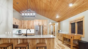 A modern kitchen with wooden cabinets, a stainless steel refrigerator, a large island with stools, and a wooden ceiling.