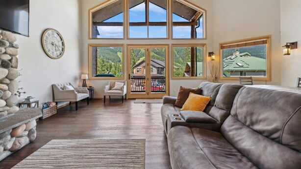 A spacious living room with a stone wall, a large gray sofa, and multiple windows offering views of a mountainous landscape. - Ski-in/Ski-Out with Private Hot Tub (Whitefish)