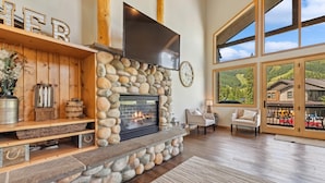 A spacious living room with a stone fireplace, wooden shelves, and a large window offering a scenic view. - Ski-in/Ski-Out with Private Hot Tub (Whitefish)