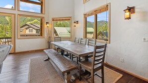 A dining area with a wooden table and benches, large windows with views of a mountain landscape, and a vase with branches on the wall. - Ski-in/Ski-Out with Private Hot Tub (Whitefish)