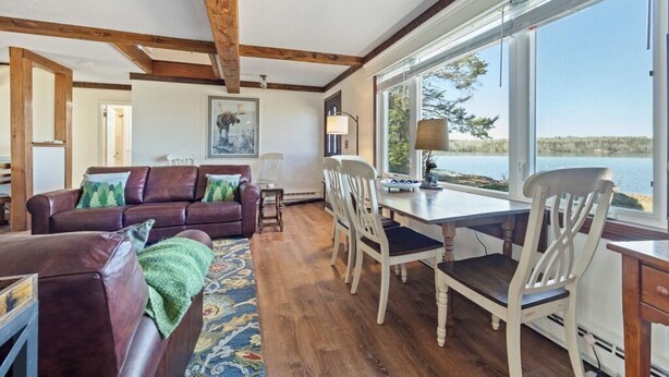 Ein Wohnzimmer mit braunem Ledersofa, weißen Stühlen, einem Holzesstisch und großen Fenstern mit Blick auf ein Gewässer.