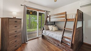Een slaapkamer met een houten stapelbed, een dressoir en een schuifdeur die uitkomt op een bebost gebied.