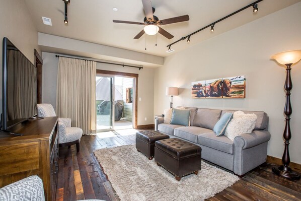 A spacious living room with a grey sofa, a wooden coffee table, and a television. There is a sliding glass door leading to an outdoor area. - Private Balcony with Outdoor Fire Table (Whitefish)