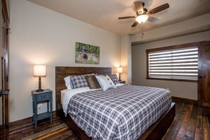 A bedroom with a wooden bed, two bedside tables with lamps, a ceiling fan, a window with blinds, and a painting on the wall. - Private Balcony with Outdoor Fire Table (Whitefish)
