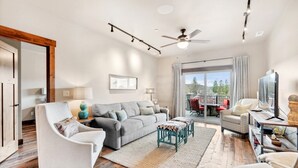 A spacious living room with a grey sofa, white armchairs, and a teal ottoman. A ceiling fan and recessed lighting are visible. - Penthouse with Hot Tub and Fire Table (Whitefish)