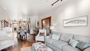 A spacious living room with a grey sofa, a matching ottoman, and a dining area with a wooden table and chairs. - Penthouse with Hot Tub and Fire Table (Whitefish)