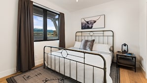 A bedroom with a metal bed frame, a view of mountains through a window, a mounted picture of a bull, a bedside table with a lantern, and a rug with geometric patterns.