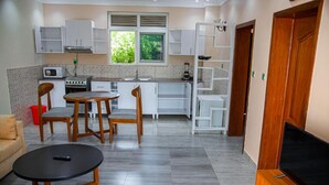 Cozinha moderna com armários brancos, sala de jantar com cadeiras de madeira, mesa redonda preta e TV fixada na parede.