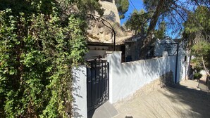 Una casa de piedra con entrada protegida por una reja, rodeada de vegetación y un cielo despejado.