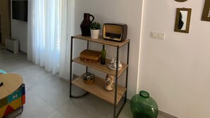 Una habitación con un estante de madera que sostiene una radio, una planta y una caja. Hay un jarrón verde en el suelo y una mesa colorida con una 'H'.