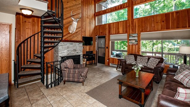 Un salón espacioso con chimenea, pared de piedra, escalera de madera y grandes ventanas que ofrecen vistas al exterior.
