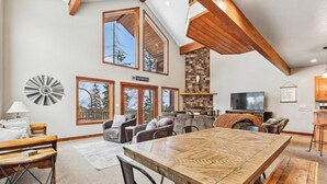 Ein geräumiges Wohnzimmer mit großem Holzesstisch, bequemen Sitzgelegenheiten, einem Kamin und großen Fenstern mit Blick ins Freie.