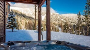 Ein Whirlpool mit Holzkonstruktion, umgeben von schneebedeckten Bäumen und Bergen.