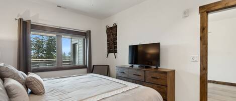 Une chambre à coucher avec un lit, un ventilateur de plafond, un téléviseur posé sur une commode en bois et une fenêtre donnant sur des arbres.