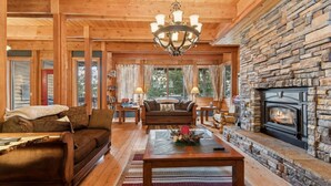 A cozy log cabin interior with a stone fireplace, wooden furniture, and large windows. - 5 Mins to Whitefish Resort and Amenities (Whitefish)