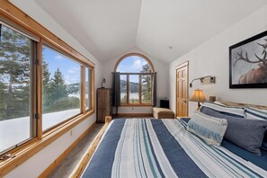 A bedroom with a large bed, a view of trees and mountains through large windows, and a mounted deer picture on the wall. - Charming Vacation Home off Big Mountain Road! Firepit! (Whitefish)