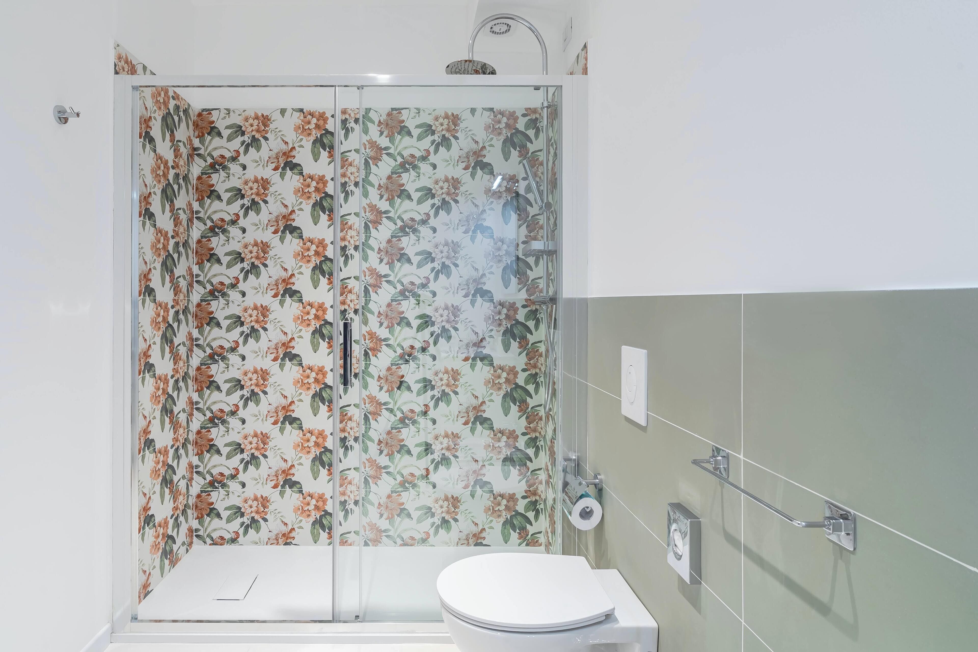Bagno con box doccia, carta da parati a motivi floreali, vaso igienico bianco e mancorrente.