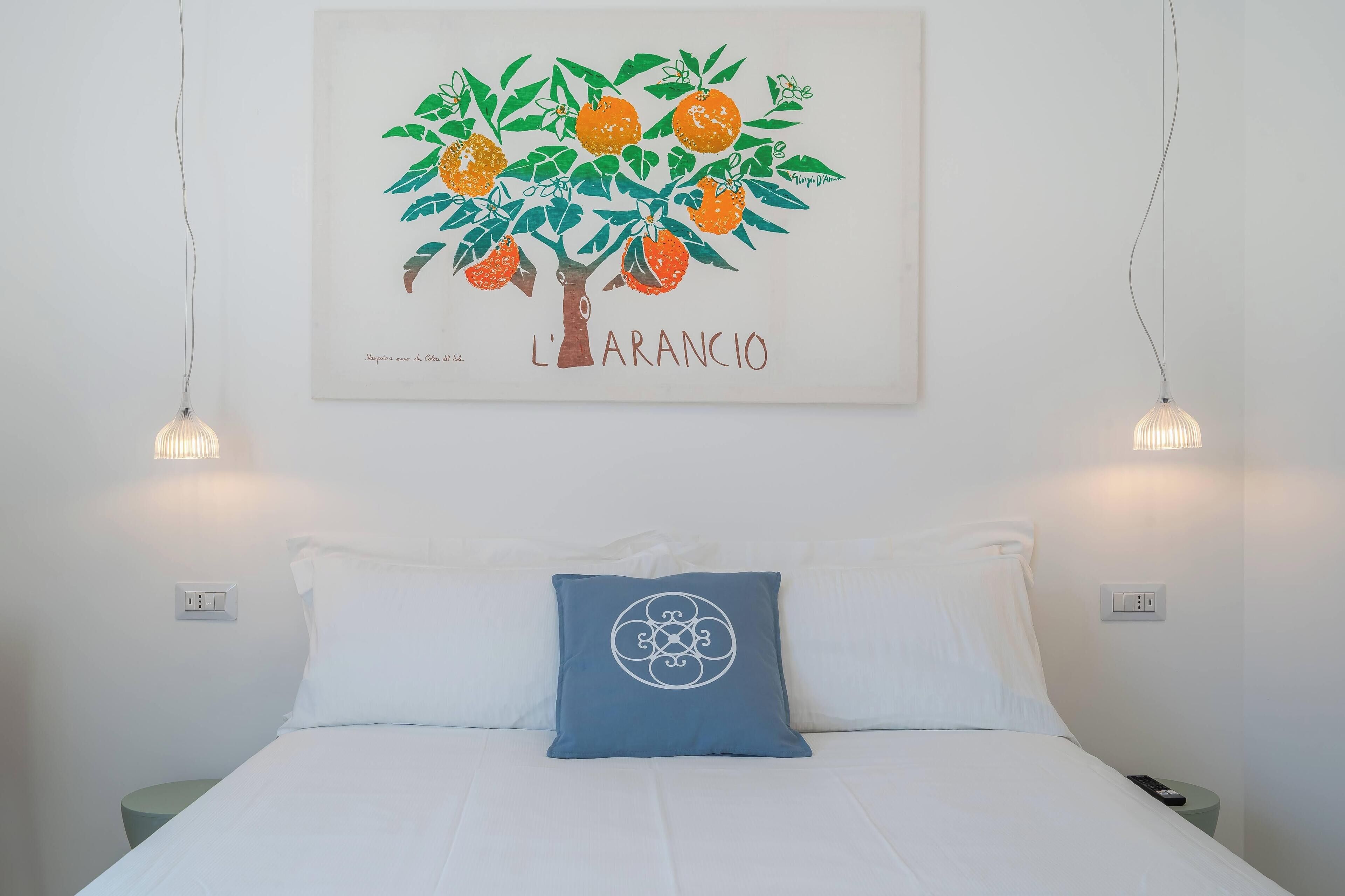 A bedroom with a bed, a pillow, a wall-mounted painting of an orange tree, and two wall-mounted lights.