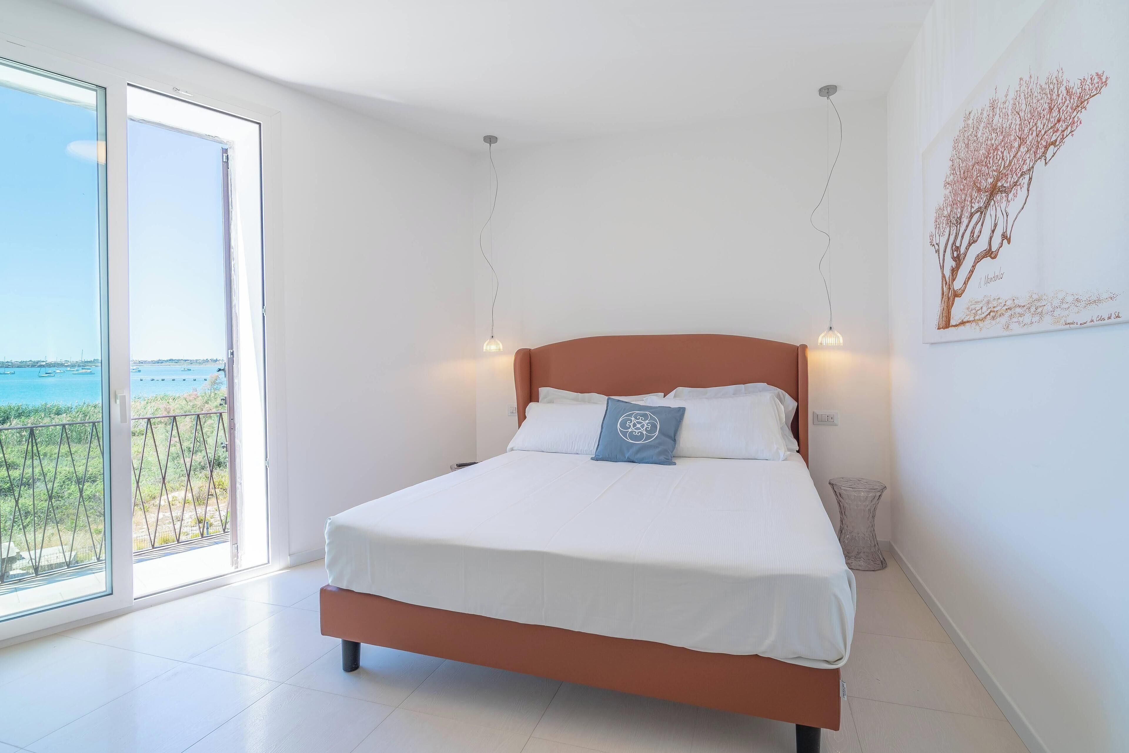 A bedroom with a bed, a view of the sea, and a painting on the wall.
