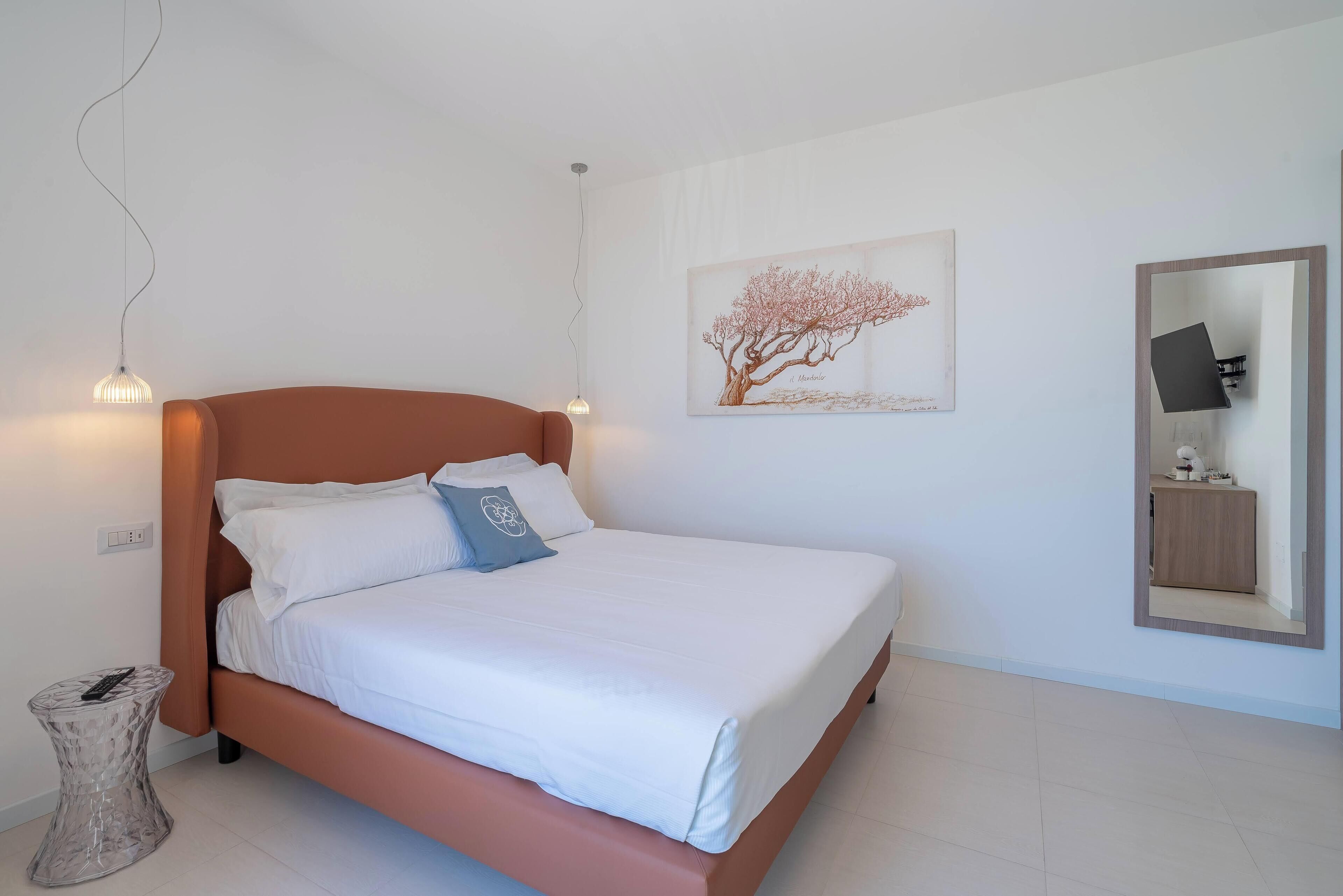 Una cama bien hecha con sábanas blancas y una única almohada decorativa, una obra de arte en la pared con un árbol, y un espejo que refleja parte de la habitación.