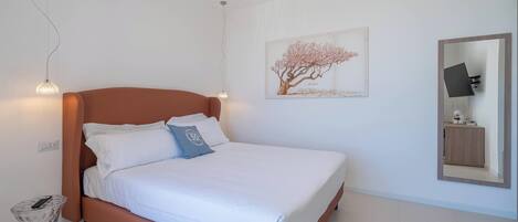 Una cama bien hecha con sábanas blancas y una única almohada decorativa, una obra de arte en la pared con un árbol, y un espejo que refleja parte de la habitación.