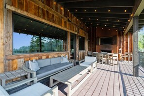 Un espace extérieur aménagé, abrité et doté de bancs et de tables en bois, d’une cheminée et de grandes fenêtres donnant sur une zone boisée.
