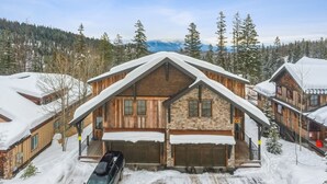 A log cabin with a stone foundation and wooden exterior, surrounded by snow-covered trees and a mountainous backdrop.