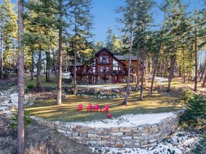 A log cabin with a stone foundation, surrounded by a stone wall and a snow-covered yard. - Home on Lost Loon Lake! Minutes to Downtown Whitefish! (Whitefish)