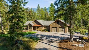 A two-story log cabin with a gravel driveway and a covered porch, surrounded by dense forest and mountains in the background. - Mountain Home with Private Hot Tub (Columbia Falls)