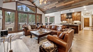 A spacious living area with leather sofas, a wooden coffee table, and a large window offering a view of the forest. - Mountain Home with Private Hot Tub (Columbia Falls)