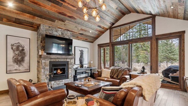 A spacious living room with a stone fireplace, leather sofas, and large windows offering a view of the outdoors. - Mountain Home with Private Hot Tub (Columbia Falls)