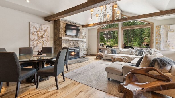 A spacious living room with a fireplace, wooden beams, a dining area, and large windows offering a view of the outdoors. - Ski-in/Ski-out Access and Private Hot Tub (Whitefish)