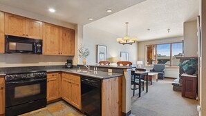 A modern kitchen with wooden cabinets, a built-in microwave, and a stove. There is a dining area with a table and chairs, and a view of the outside through a window.