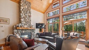 A cozy living room with a stone fireplace, leather furniture, and large windows offering a view of the forest.