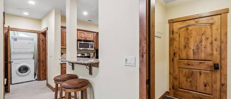A compact kitchen with a washing machine, a microwave, and wooden bar stools.