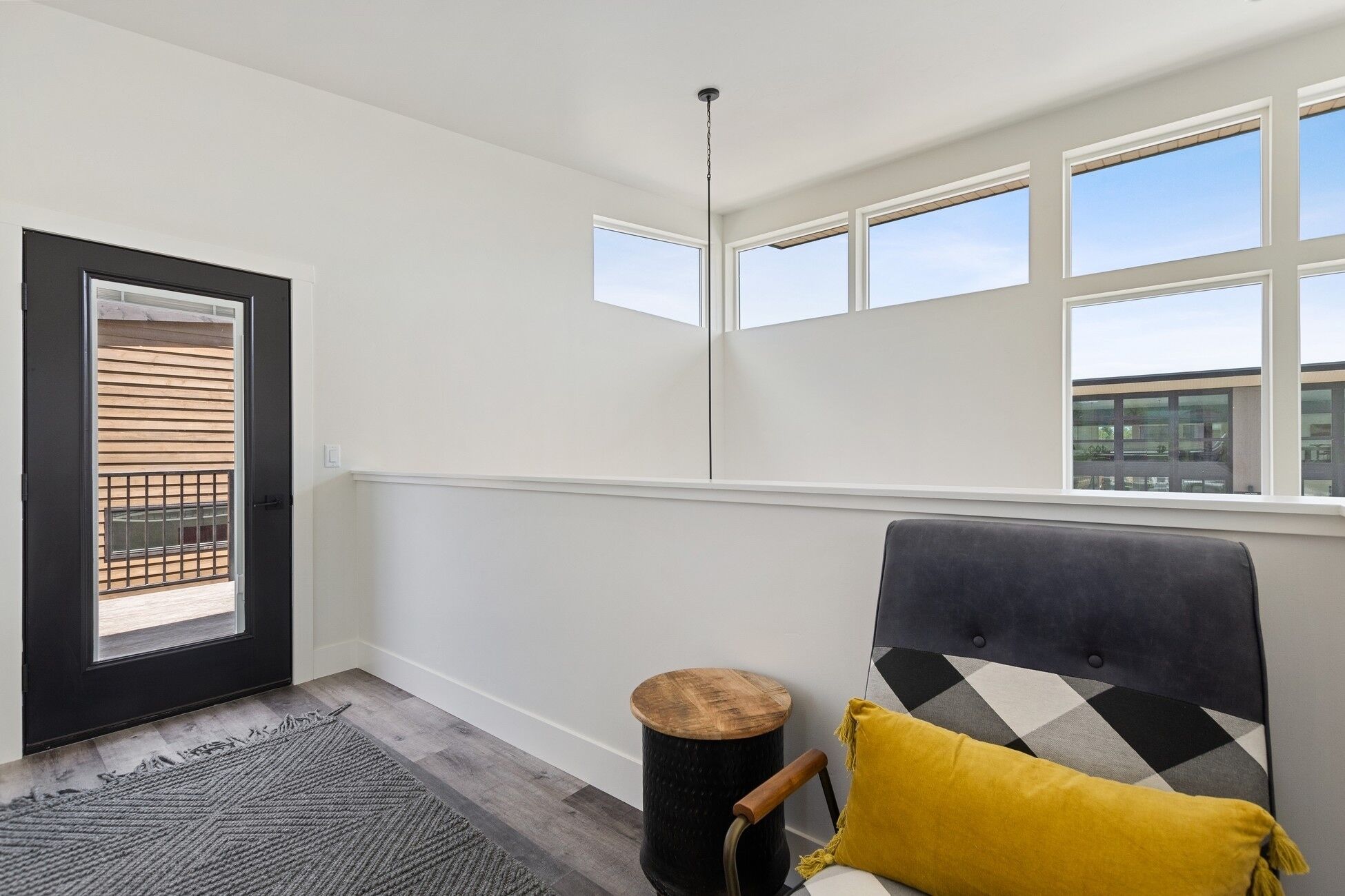 A modern room with a large window, a door with a wooden shutter, a round wooden stool, and a grey upholstered chair with a yellow cushion.