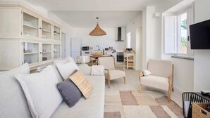 Amplia sala de estar con sofá, sillas, una cocina al fondo y un televisor montado en la pared.