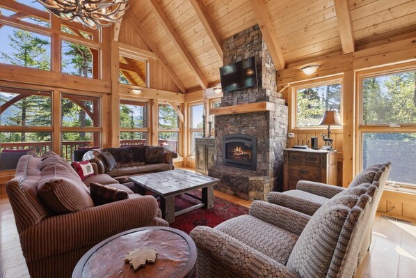A cozy cabin interior with a stone fireplace, wooden beams, and large windows offering views of the forest.