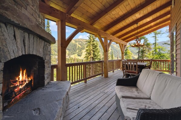 Una terraza de madera con chimenea de piedra, mobiliario exterior y vistas a un paisaje montañoso.
