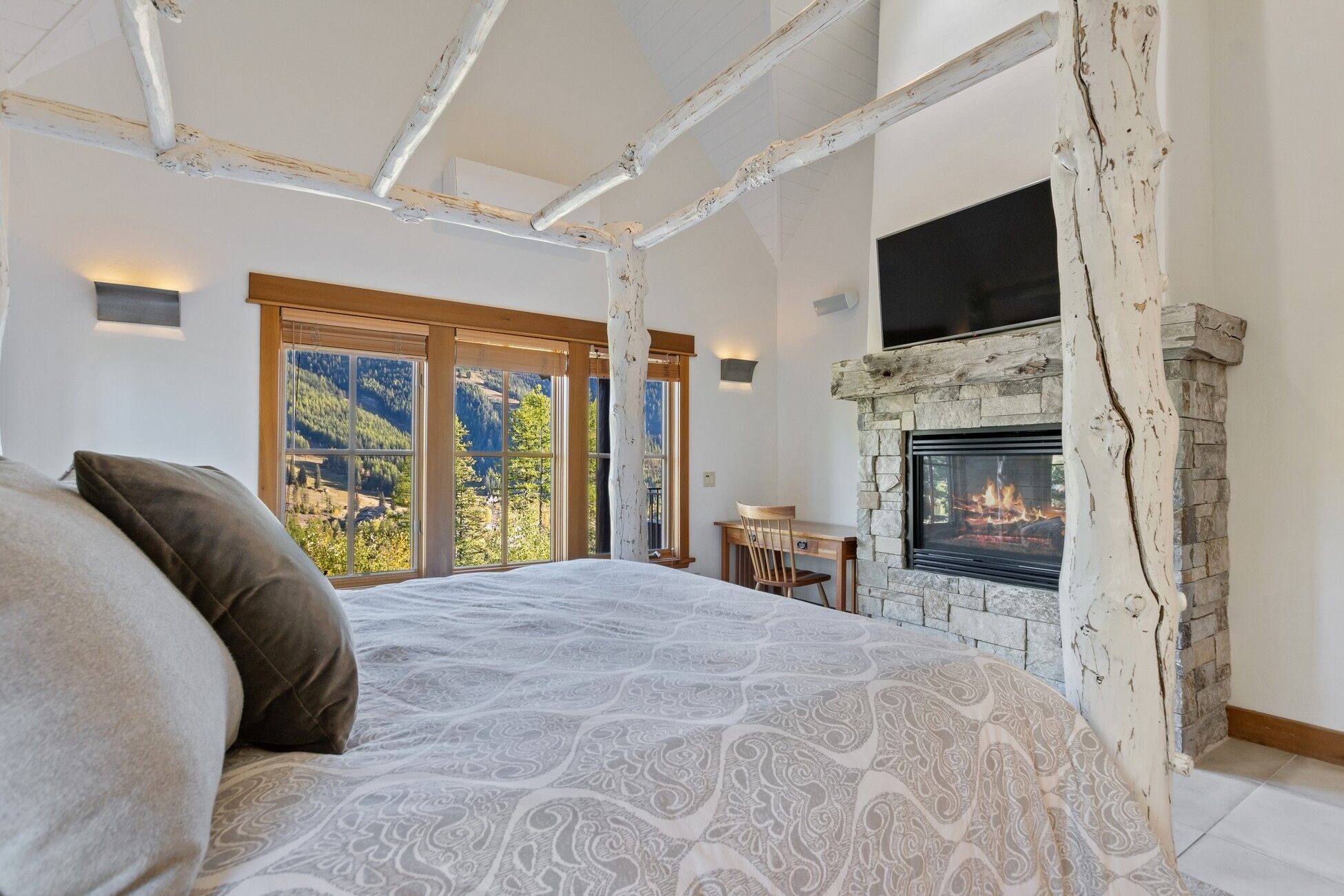 Une chambre dotée d’un grand lit, d’une cheminée en pierre et offrant une vue sur l’extérieur grâce à de grandes fenêtres.