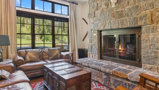 Un salón acogedor con chimenea de piedra, sofá de cuero y grandes ventanales con vistas a un bosque.