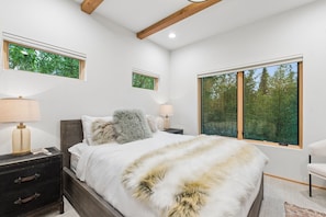 A bedroom with a bed, two bedside tables, a nightstand, a chair, and a large window with a view of trees. - Boot Dryers | Ski-in/out | Hot Tub (Whitefish)