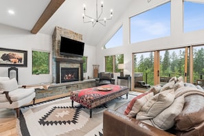 A spacious living room with a stone fireplace, leather furniture, and large windows offering a view of the forest.