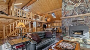 A log cabin interior with a stone fireplace, a leather sofa, a dining table, and a staircase leading to an upper level.
