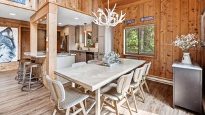 L’intérieur d’un chalet en bois comprend une grande table à manger, quatre chaises, un lustre et un coin cuisine.